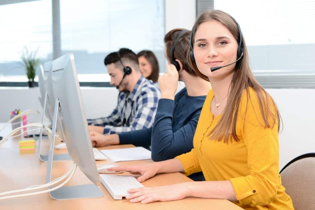 portrait of beautiful and cheerful young woman telephone operator with headset working on desktop computer in row in a customer service call support helpline business center with teamworker in background