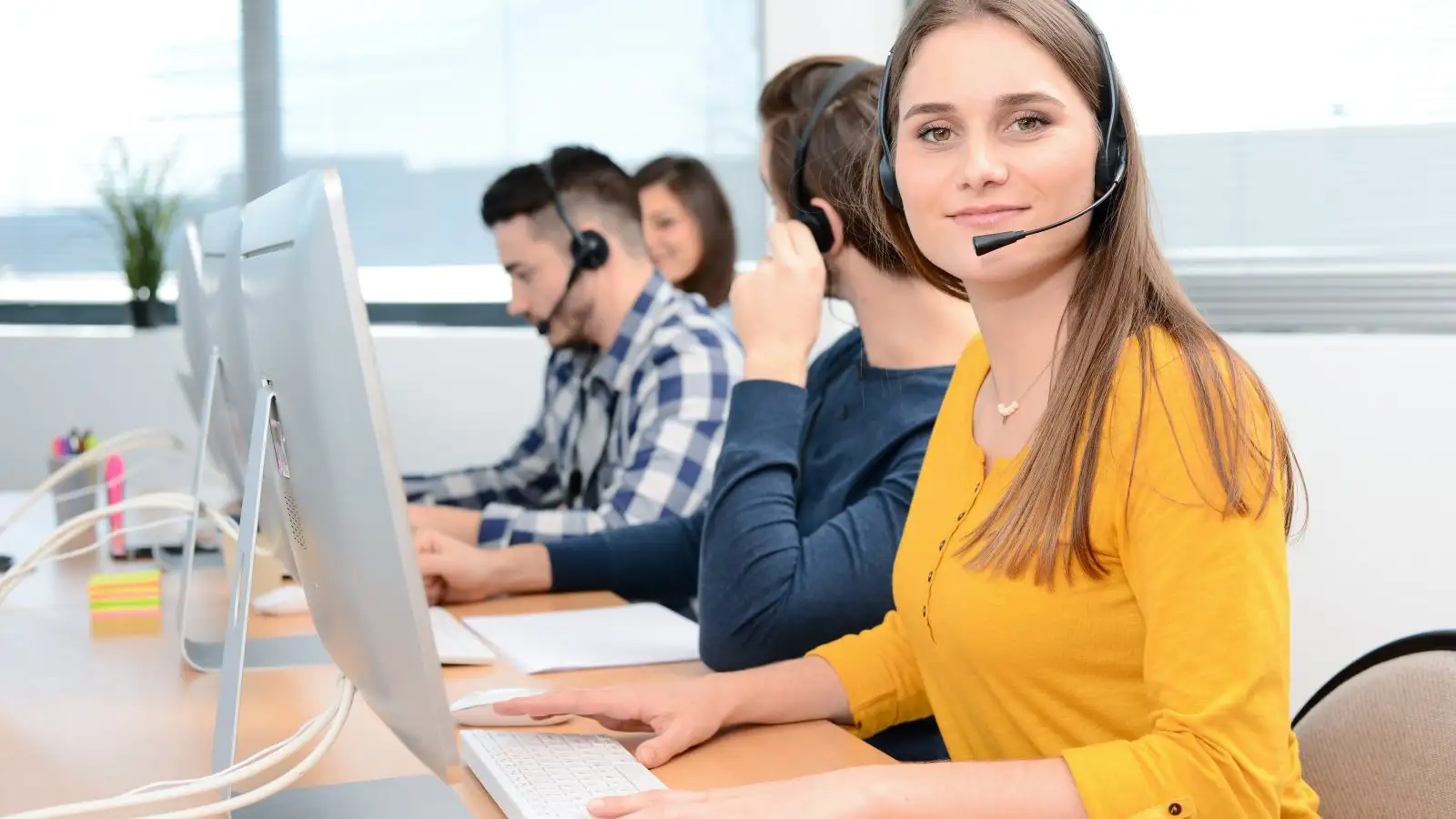 portrait of beautiful and cheerful young woman telephone operator with headset working on desktop computer in row in a customer service call support helpline business center with teamworker in background
